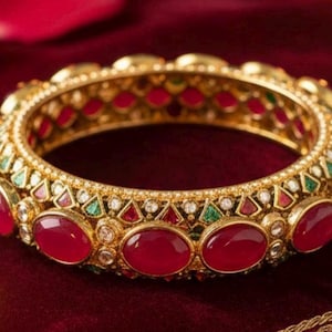 May include: A gold-colored bangle bracelet with red, green, and white gemstones. The bracelet features oval-shaped red stones and smaller accent stones. The jewelry is displayed on a dark red velvet surface.