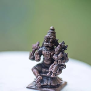 May include: A bronze-colored statue of a deity holding a smaller figure. The larger figure has a detailed face with a mustache and a tall headdress. The smaller figure is also adorned with a headdress. The statue is on a square base.