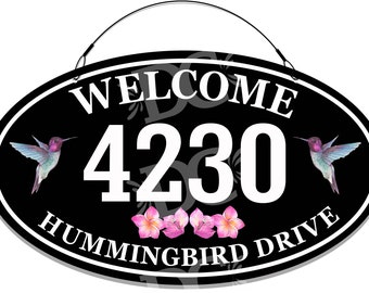 May include: Black oval welcome sign with white text that reads "Welcome 4230 Hummingbird Drive". The sign has two hummingbird illustrations and pink flowers.