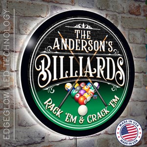 May include: A round sign with a black and green design, featuring a white border and a lit edge. The sign reads "The Anderson's Billiards Rack 'Em & Crack 'Em" with crossed pool cues and billiard balls on a green felt table. The sign is made in the USA.