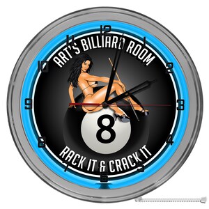 May include: A blue neon clock with a black background featuring a woman sitting on a white billiard ball with the number 8. The clock face reads "Art's Billiard Room" and "Rack It & Crack It".