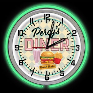 May include: A circular wall clock with a neon green surround. The clock face displays "Percy's DINER" in pink and black, alongside illustrations of a milkshake, burger, and chips. The words "Good Eats!" are also visible.