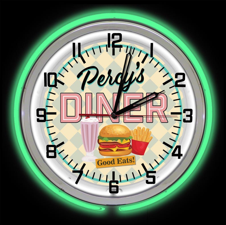 May include: A circular wall clock with a neon green surround. The clock face displays "Percy's DINER" in pink and black, alongside illustrations of a milkshake, burger, and chips. The words "Good Eats!" are also visible.
