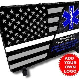 Könnte beinhalten: Schwarz-weiße amerikanische Flagge mit einem dünnen weißen Streifen in der Mitte. Die Flagge befindet sich auf einem schwarzen Schiefer mit einem blauen Lebensstern und einer weißen Schlange. Der Text lautet "PARAMEDIC TYLER MITCHELL OKTOBER 1999 - FEBRUAR 2025 26 JAHRE TREUE UND HINGABE GREENWOOD VILLAGE EMS".