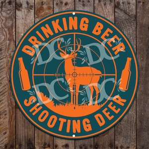 May include: A round, teal-colored metal sign with the text "Drinking Beer Shooting Deer" in orange letters. The sign features a deer silhouette in the center of a target with two orange beer bottles on either side.