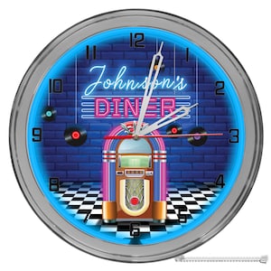 May include: A silver framed clock with a blue neon ring around the edge. The clock face features a black and white checkered floor, a brick wall with a jukebox, and a pink neon sign that reads "Johnson's Diner".