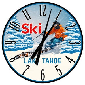 Può includere: Un orologio rotondo con uno sfondo blu e bianco che presenta uno sciatore con una giacca arancione e pantaloni blu. Il quadrante dell'orologio ha numeri romani e le parole "Ski Lake Tahoe" in rosso e blu.