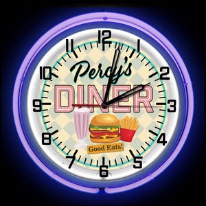 May include: Circular wall clock with a neon blue surround. The clock face displays "Percy's DINER" in pink and black, with illustrations of a milkshake, burger, and fries. The words "Good Eats!" are also visible.