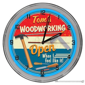 May include: A blue and red vintage-style clock with a silver frame. The clock face says "Tom's Woodworking Open When I Feel Like It!" and features a hammer, a ruler, and other woodworking tools.