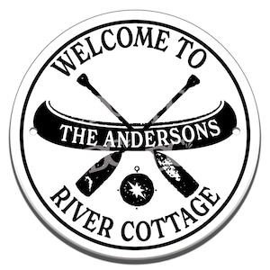 Puede incluir: Señal redonda en blanco y negro con el texto "Bienvenido a The Andersons River Cottage" y un gráfico de dos remos de canoa cruzados con una brújula en el centro.