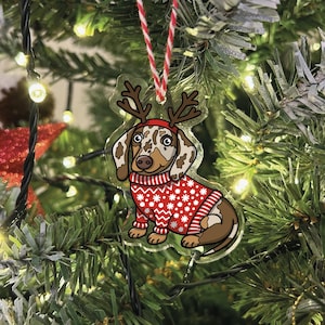 Può includere: Un ornamento di Natale in plastica trasparente con un bassotto dei cartoni animati che indossa un maglione rosso e bianco e corna di renna.