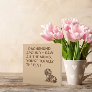 May include: A card with a brown background and a cartoon dachshund. The card reads "I dachshund around + saw all the mums. You're totally the best!" The card is sitting on a wooden table with a bouquet of pink tulips in a white mug with a heart pattern.
