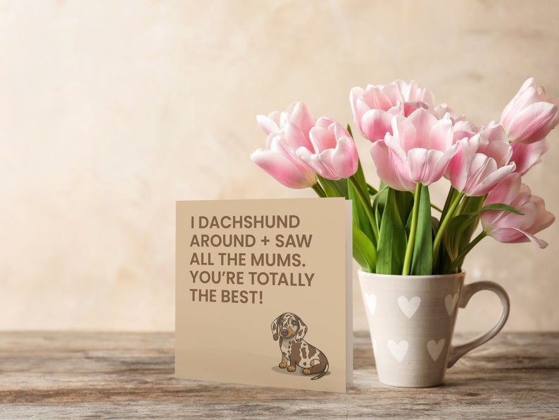 May include: A card with a brown background and a cartoon dachshund. The card reads "I dachshund around + saw all the mums. You're totally the best!" The card is sitting on a wooden table with a bouquet of pink tulips in a white mug with a heart pattern.