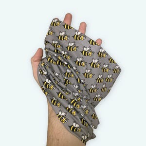 May include: A gray neck gaiter with a repeating pattern of yellow and black cartoon bees. The bees have smiling faces and are flying.