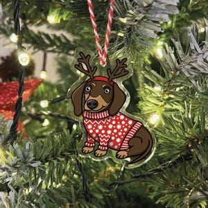May include: A brown and tan dachshund wearing a red and white Christmas sweater and reindeer antlers. The dachshund is a Christmas ornament made of clear acrylic.