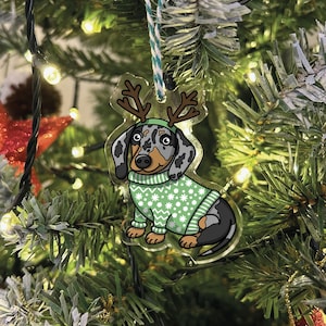 May include: A Christmas ornament featuring a cartoon dachshund wearing a green sweater with a snowflake pattern and reindeer antlers. The dog has a black and grey spotted coat.