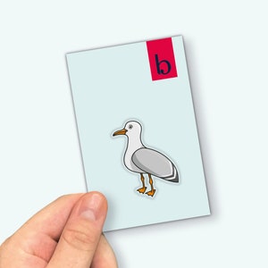 May include: A sticker featuring a cartoon illustration of a white seagull with orange feet and a grey wing. The sticker is on a light blue background with a red rectangle with the letter "b" in black.