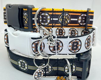 Puede incluir: Tres collares para perros con un patrón de logotipo de los Boston Bruins en negro, blanco y amarillo. Los collares están superpuestos y tienen anillos y dijes de metal plateado.