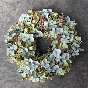 Könnte beinhalten: Ein kreisförmiger Kranz aus getrockneten Hortensienblüten in staubigen Blau- und Salbeigrüntönen, durchsetzt mit kleinen dunkelvioletten Knospen, getrocknetem Laub und zarten weißen Füllblumen, der ein dichtes und strukturiertes botanisches Arrangement bildet.