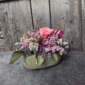 Könnte beinhalten: Ein Blumenarrangement mit einer zentralen pfirsichfarbenen Rose, umgeben von violetten, rosafarbenen und weißen Trockenblumen sowie Grünpflanzen. Das Arrangement befindet sich in einem ovalen, hellgrünen Keramikübertopf mit strukturierter, gerippter Oberfläche.
