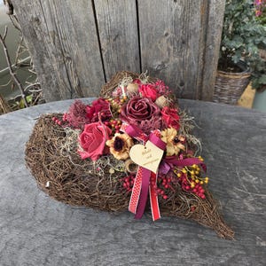 Könnte beinhalten: Ein herzförmiger Kranz aus braunen geflochtenen Zweigen, verziert mit dunkelroten und rosafarbenen künstlichen Rosen, Trockenblumen und roten Beeren. Ein kleines hölzernes Herzschild mit der Aufschrift "Geliebt und unvergessen" ist mit einem rot-weiß gepunkteten Band befestigt.