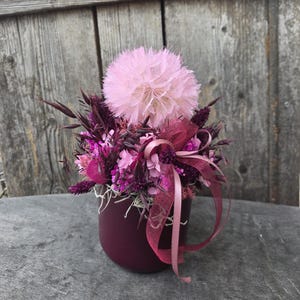 Könnte beinhalten: Ein Blumenarrangement in einem dunkel-kastanienbraunen zylindrischen Gefäß mit einer großen, flauschigen, hellrosa, löwenzahnartigen Blüte, umgeben von dunkelvioletten Trockengräsern, leuchtend rosa Blüten und einer halbtransparenten, kastanienbraunen Schleife.