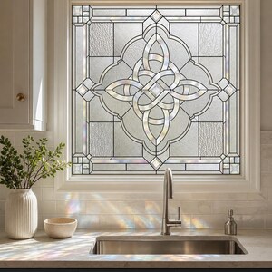 May include: A square window featuring decorative leaded glass with a central intricate knot pattern. The glass panels include clear, textured, and beveled sections that create light refractions. The window is framed in white wood trim.