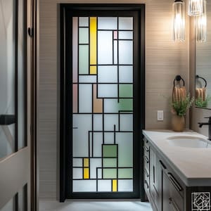 May include: A bathroom with a black framed door with a frosted glass panel featuring a geometric design in yellow, green, pink, and white. The door is in the center of the image. The bathroom has a white countertop with a sink and a wooden cabinet with drawers.