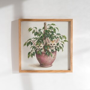 Op de afbeelding: Een vierkante ingelijste muurkunstprint met een schilderij van twee woestijnvossen met grote oren die achter de groene bladeren van een kamerplant vandaan komen. De plant staat in een roze pot met textuur, gevat in een lichthouten lijst.