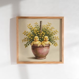 May include: A square framed wall art piece featuring a painting of two yellow ducklings sitting inside a terracotta-colored flower pot. The pot is filled with a lush green and yellow leafy plant, and the entire image is encased in a light wood frame.