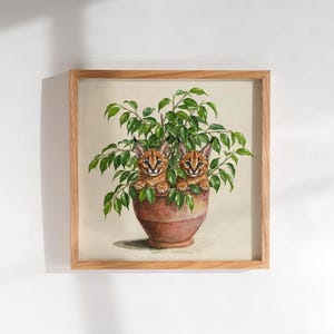May include: A square framed wall art print featuring a painting of two caracal kittens peeking out from a lush green leafy plant potted in a terracotta-colored clay pot. The artwork is encased in a light-toned wooden frame with a clean finish.