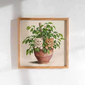 May include: A square framed wall art piece featuring a painting of two small kittens, one cream and one orange tabby, peeking out from a lush green ficus plant potted in a terracotta-colored clay pot. The frame is made of light-toned wood.