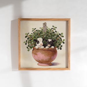 May include: A square framed wall art print featuring a painting of two small calves, one black and white and one solid black, peeking out from a large terracotta-colored pot filled with a lush green leafy plant with a wooden stake in the center.