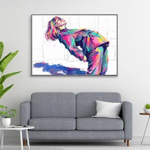 Peut inclure: Une impression d'art pop encadrée d'une personne en costume coloré, penchée en arrière. L'œuvre présente des teintes vibrantes de rose, de bleu, de jaune et de vert. L'impression est affichée au-dessus d'un canapé gris, avec un oreiller blanc et une petite table d'appoint.