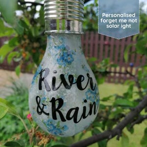 Op de afbeelding: Een decoratieve gloeilamp met een zilveren voet en een matglazen behuizing. De lamp is versierd met blauwe bloempatronen en de woorden "River & Rain" in zwart schrift. De tekst "Personalised forget me not solar light" is ook zichtbaar.