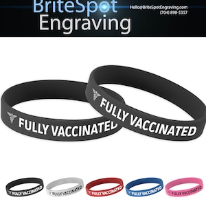 Op de afbeelding: Twee zwarte siliconen polsbandjes met de tekst "FULLY VACCINATED" en een medisch symbool. De polsbandjes worden getoond tegen een witte achtergrond.  Onder de polsbandjes bevinden zich vijf andere polsbandjes in verschillende kleuren: grijs, rood, blauw en roze.