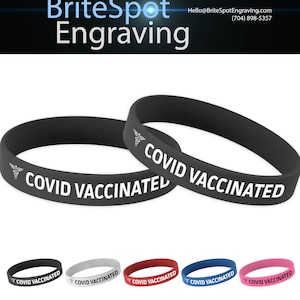 Op de afbeelding: Twee zwarte siliconen polsbandjes met de tekst "COVID VACCINATED" en een medisch symbool. De polsbandjes liggen op elkaar. Onder de polsbandjes bevinden zich zes andere polsbandjes in verschillende kleuren: zwart, grijs, rood, blauw en roze.