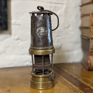 Könnte beinhalten: Eine antike Grubenlampe aus Messing und Kupfer mit Tragegriff. Die Lampe hat einen Drahtkäfig um das Glas und einen Messingfuß.