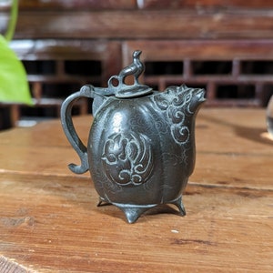 Puede incluir: Una tetera pequeña de color verde oscuro con intrincados diseños florales y una tapa con forma de pájaro. La tetera tiene cuatro patas y está sentada sobre una mesa de madera.