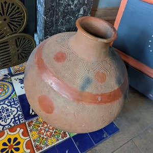 May include: A large, round, terracotta pot with a wide opening and a brown, orange, and black painted design. The pot is sitting on a tiled floor.