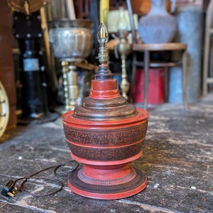 Può includere: Una lampada da tavolo decorativa rossa e nera con un design a più livelli e ornato. La lampada presenta motivi intricati e una lampadina in alto. La lampada è collegata a una presa elettrica. La lampada è su un pavimento di legno.