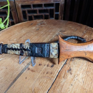 May include: A wooden-handled dagger with a black leather wrap and a bone-like handle. The dagger is on a wooden surface.