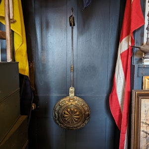 May include: A brass fireplace tool with an ornate design hangs on a dark blue wall. The tool has a long handle and a round head with a floral pattern.