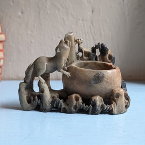 May include: A small, grey stone sculpture of a creature with a long tail and a bowl on top. The sculpture is carved with intricate details and has a rustic, antique look.