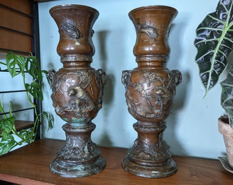 May include: Two antique bronze vases with intricate floral and bird designs. The vases have a dark brown patina and are decorated with raised details. They are tall and slender, with a wide base and a narrow neck.