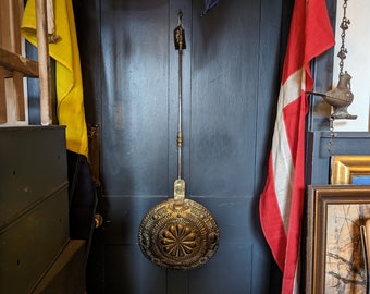May include: A brass fireplace tool with an ornate design hangs on a dark blue wall. The tool has a long handle and a round head with a floral pattern.