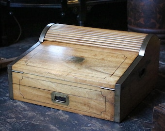 Op de afbeelding: Een vintage houten bureau met een rolluik en messing accenten. Het bureau heeft een lade met een messing handvat en is gemaakt van lichtgekleurd hout. De randen zijn versterkt met metaal. Een klassiek meubelstuk.