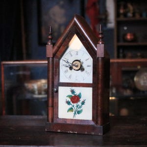 May include: Antique wooden clock with a triangular top and a white clock face featuring black Roman numerals. The clock has a decorative floral panel with a red rose and green leaves. The clock is approximately 12 inches tall.