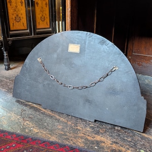 May include: A semi-circular black wooden panel featuring a metal hanging chain attached to two eyelets on the back. A small rectangular brass-colored label is affixed to the upper center of the flat surface, which shows visible wood grain texture.