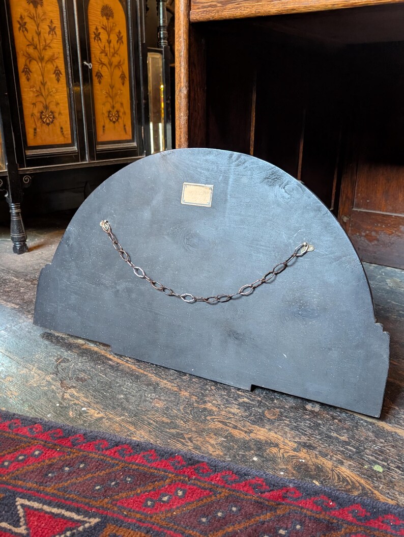 May include: A semi-circular black wooden panel featuring a metal hanging chain attached to two eyelets on the back. A small rectangular brass-colored label is affixed to the upper center of the flat surface, which shows visible wood grain texture.
